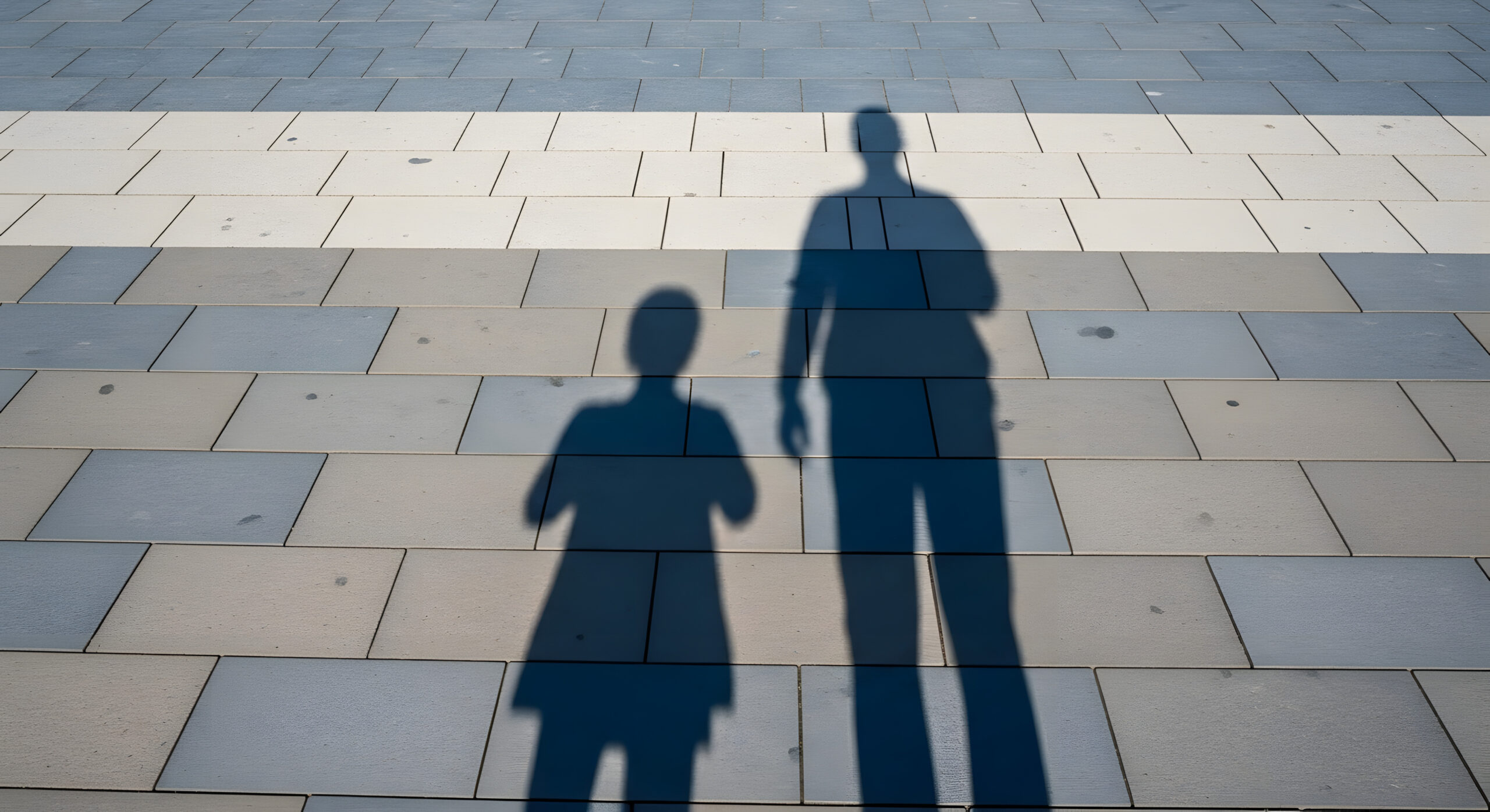 Long shadows of an adult and a child stretched across pale grey paving stones on a sunny day.