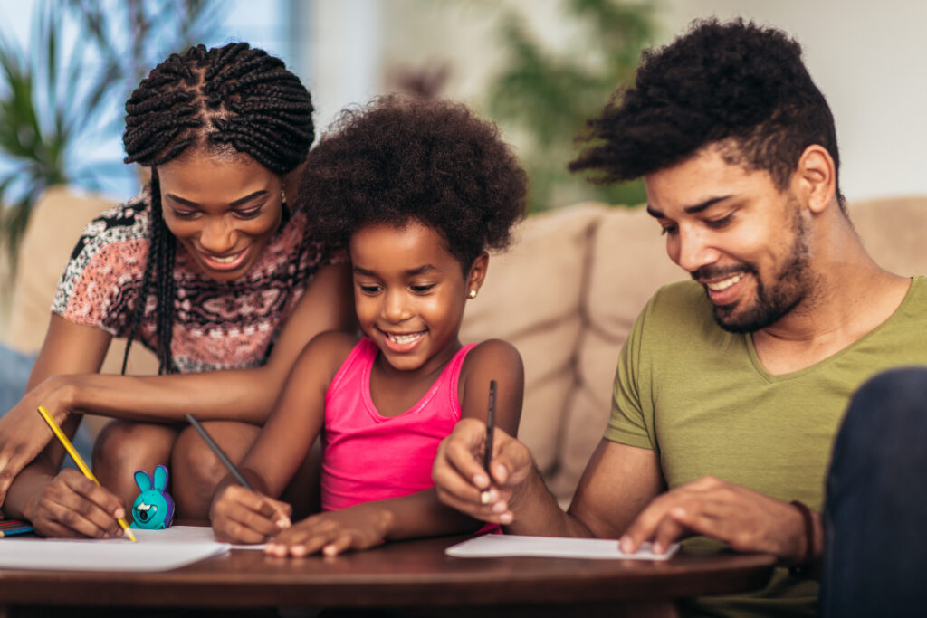 A mother and father drawing at the kitchen table with their young daughter, all three smiling at the page